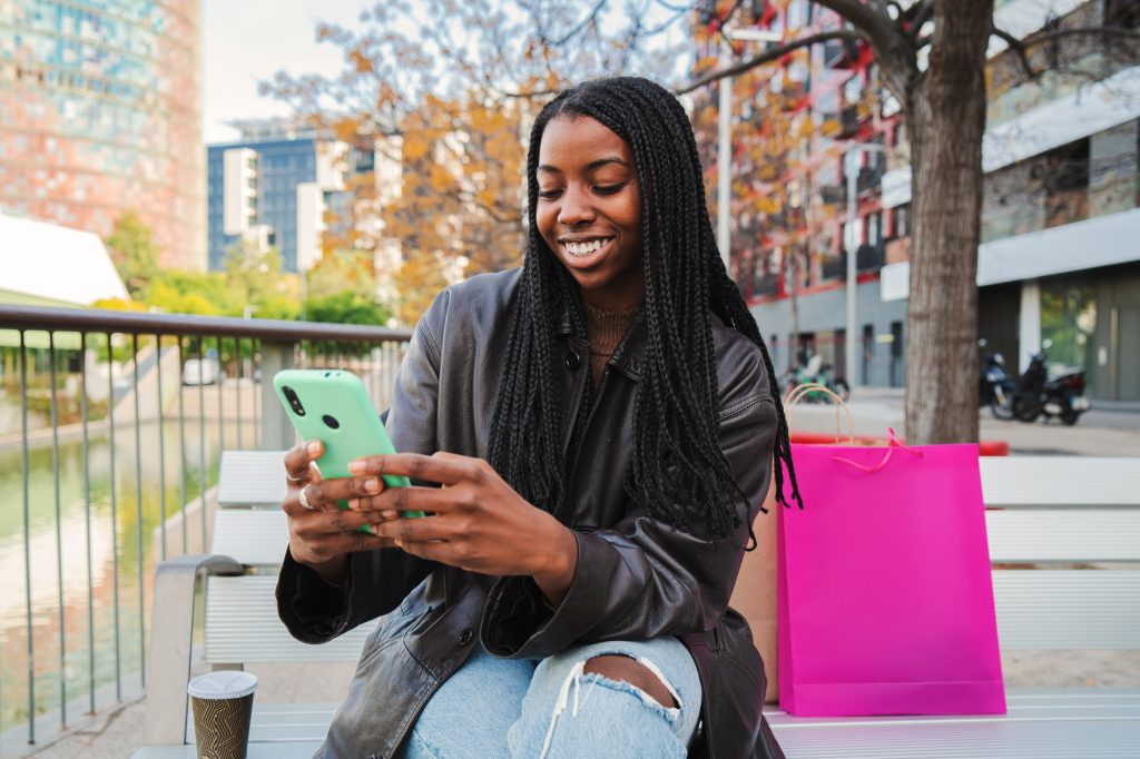 Happy teenage african american girl smiling doing shopping online with a cellphone sitting on a